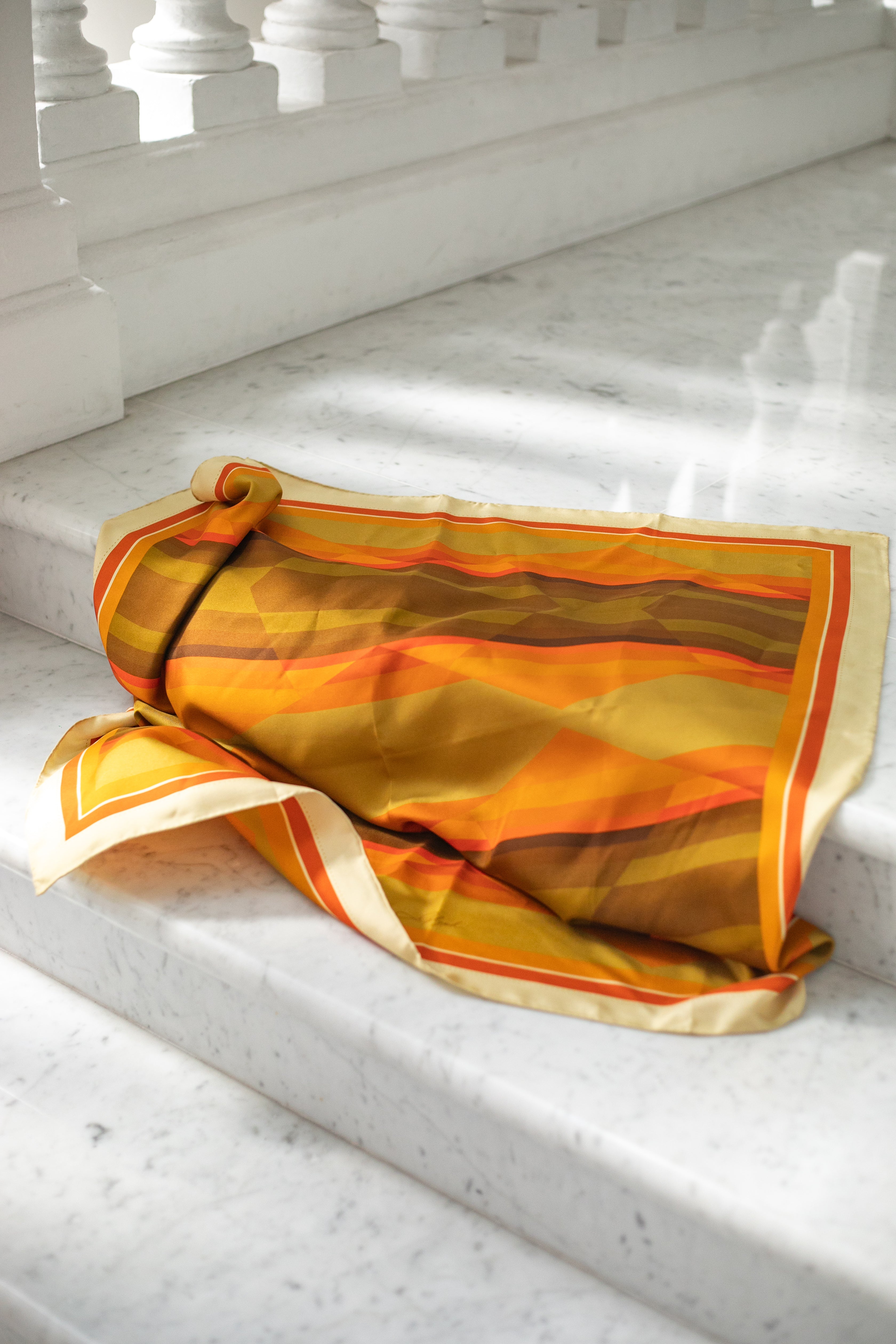 Amber Field silk scarf by Glyph draped on white marble steps, highlighting the fluid silk texture and warm amber, gold and earthy tones.