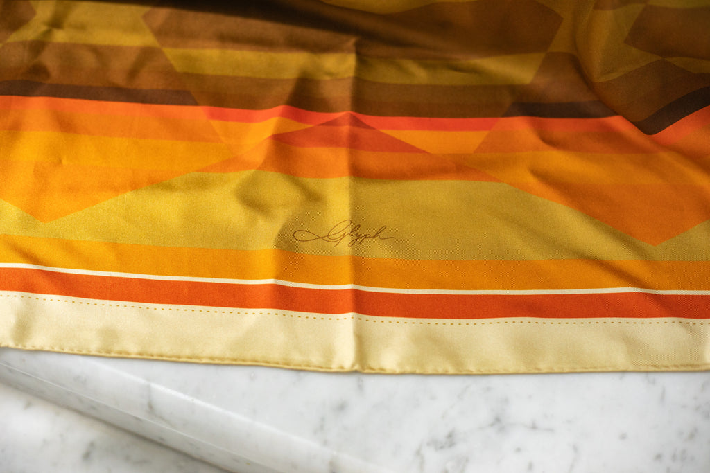 Close-up of the Amber Field silk scarf by Glyph, showing the signature, hand-rolled edge and warm amber, gold and earthy tones.