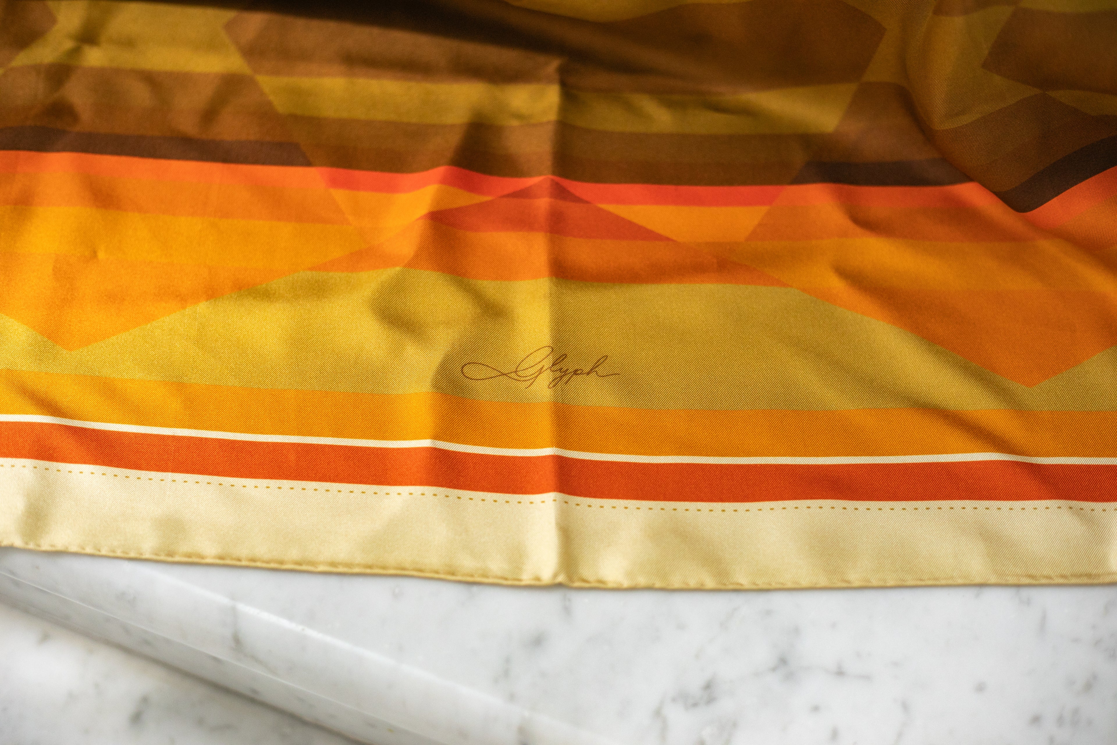 Close-up of the Amber Field silk scarf by Glyph, showing the signature, hand-rolled edge and warm amber, gold and earthy tones.