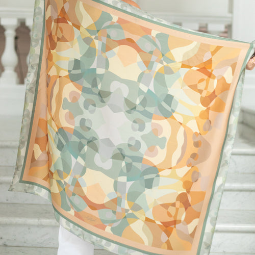 Woman holding the Harmony silk scarf by Glyph, showing the full abstract artwork in peach, sage green and soft neutral tones.